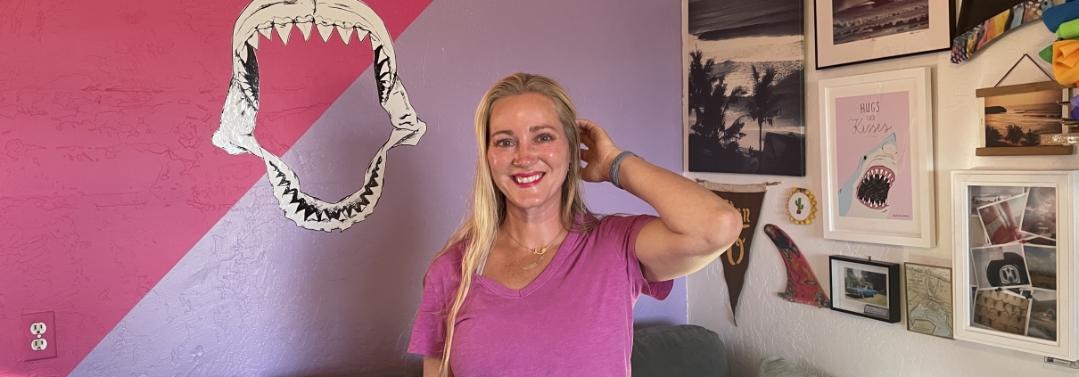 woman with long blonde hair wearing a pink shirt and white shorts smiles while standing in front of a pink wall with a shark mouth painted on the wall