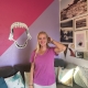 woman with long blonde hair wearing a pink shirt and white shorts smiles while standing in front of a pink wall with a shark mouth painted on the wall