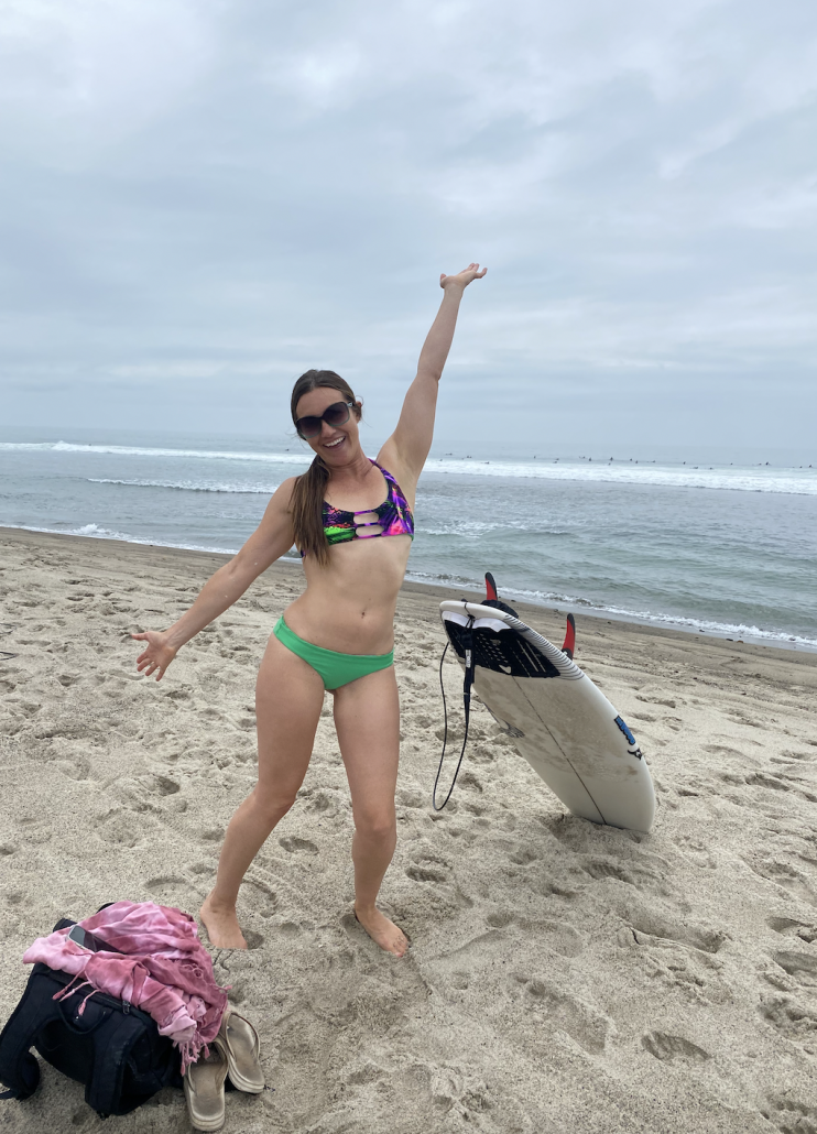 happy surfer girl standing with hands in the air on the beach in front of the ocean with her surfboard sitting in the sand